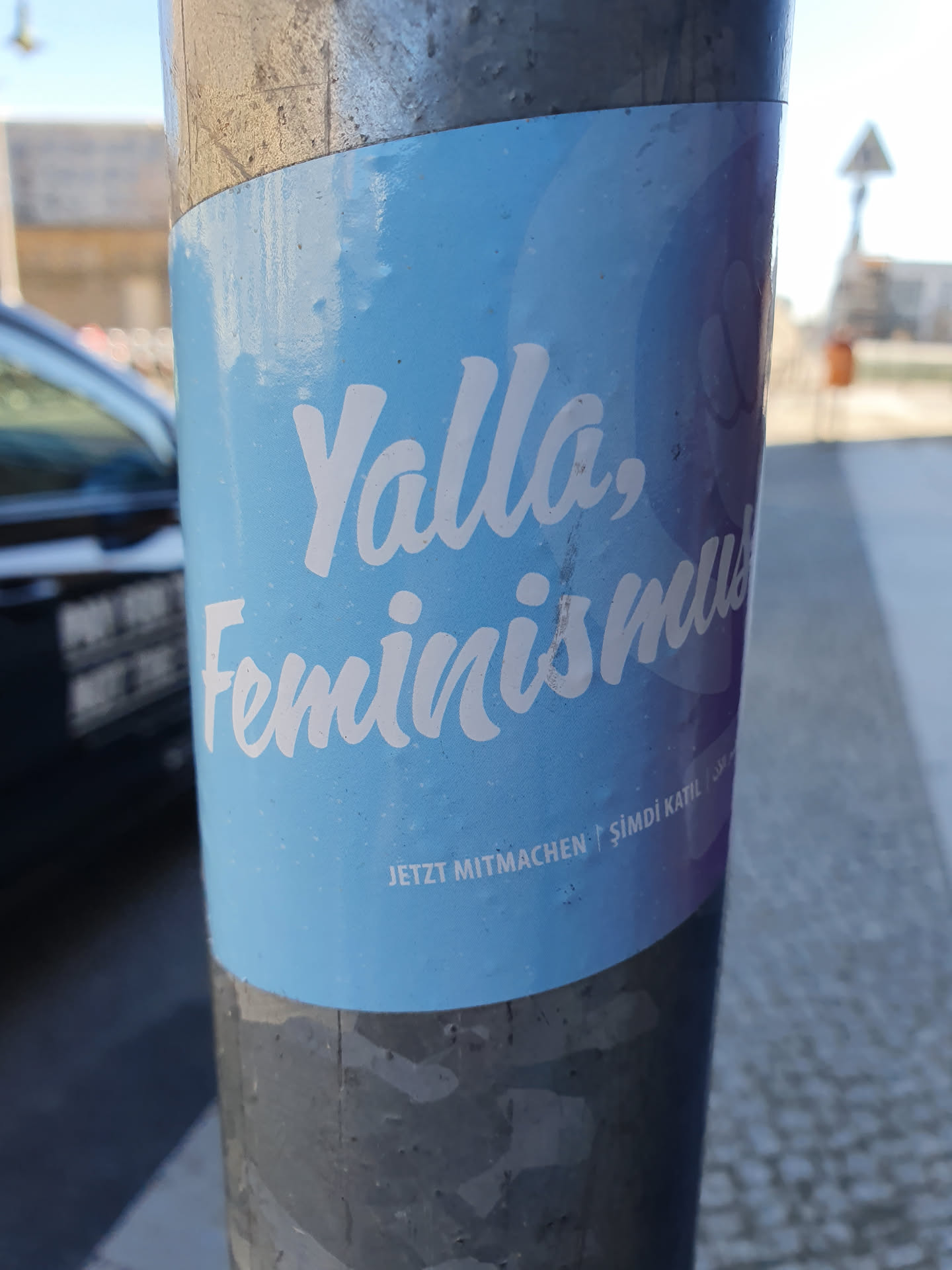 A light blue sticker on a pole with the text "Yalla, Feminismus!" in a stylized white font.  Smaller text at the bottom reads "Jetzt mitmachen!" which translates to "Join now!" in German.
