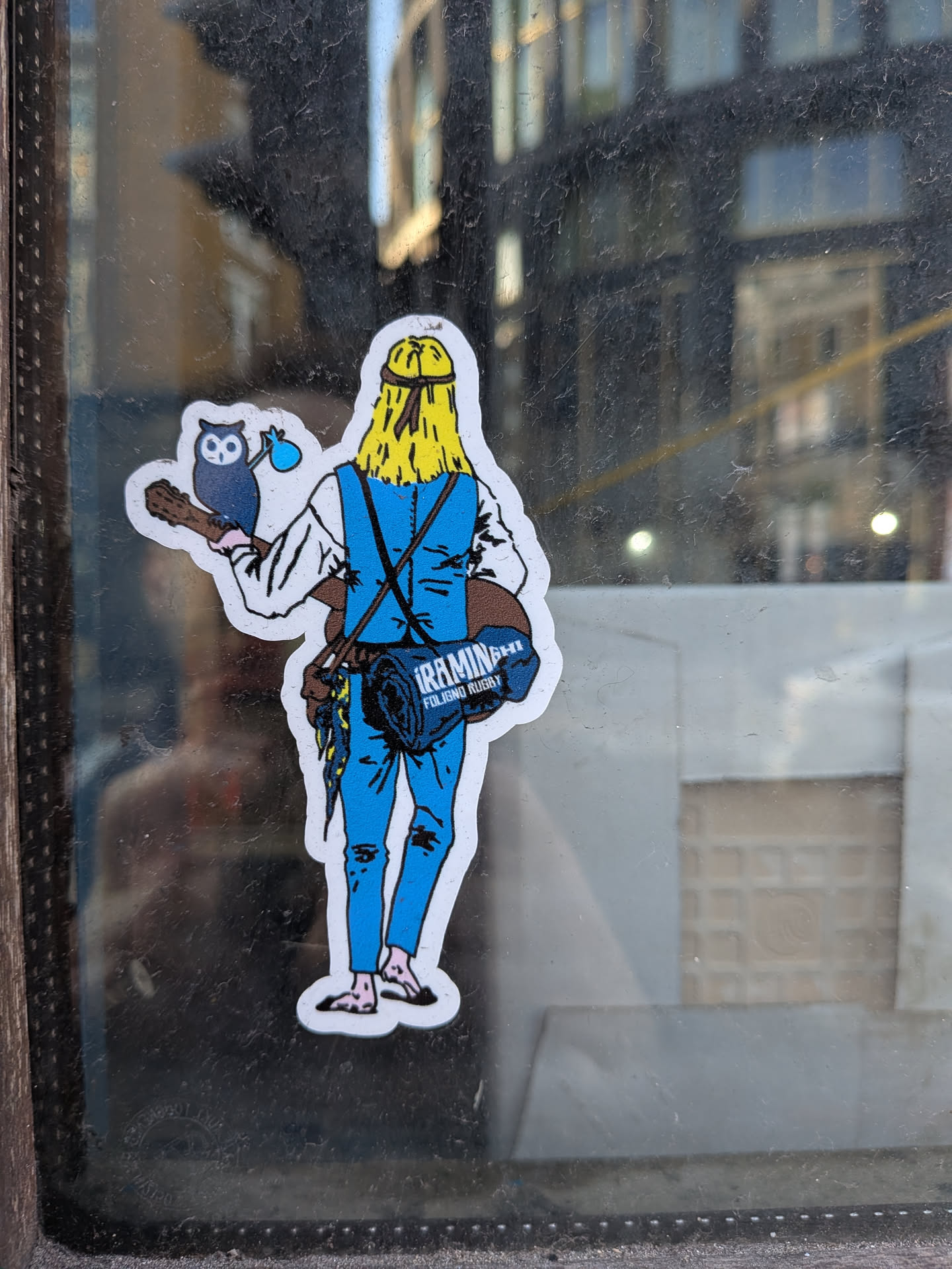 The sticker depicts a cartoon-style character viewed from the back, facing away. The character has long, bright yellow hair, tied with a dark band, and wears light blue overalls over a white long-sleeved shirt. An acoustic guitar, brown in color, is slung over their back. Perched on the guitar's neck is a small, dark blue owl holding a light blue pouch. Strapped to the character's back is a dark blue rolled-up item, possibly a sleeping bag or mat, with white text visible on it. The character is barefoot, showing light pink feet. The entire illustration is outlined with a distinct white border, suggesting it's a die-cut sticker applied to a glass surface.