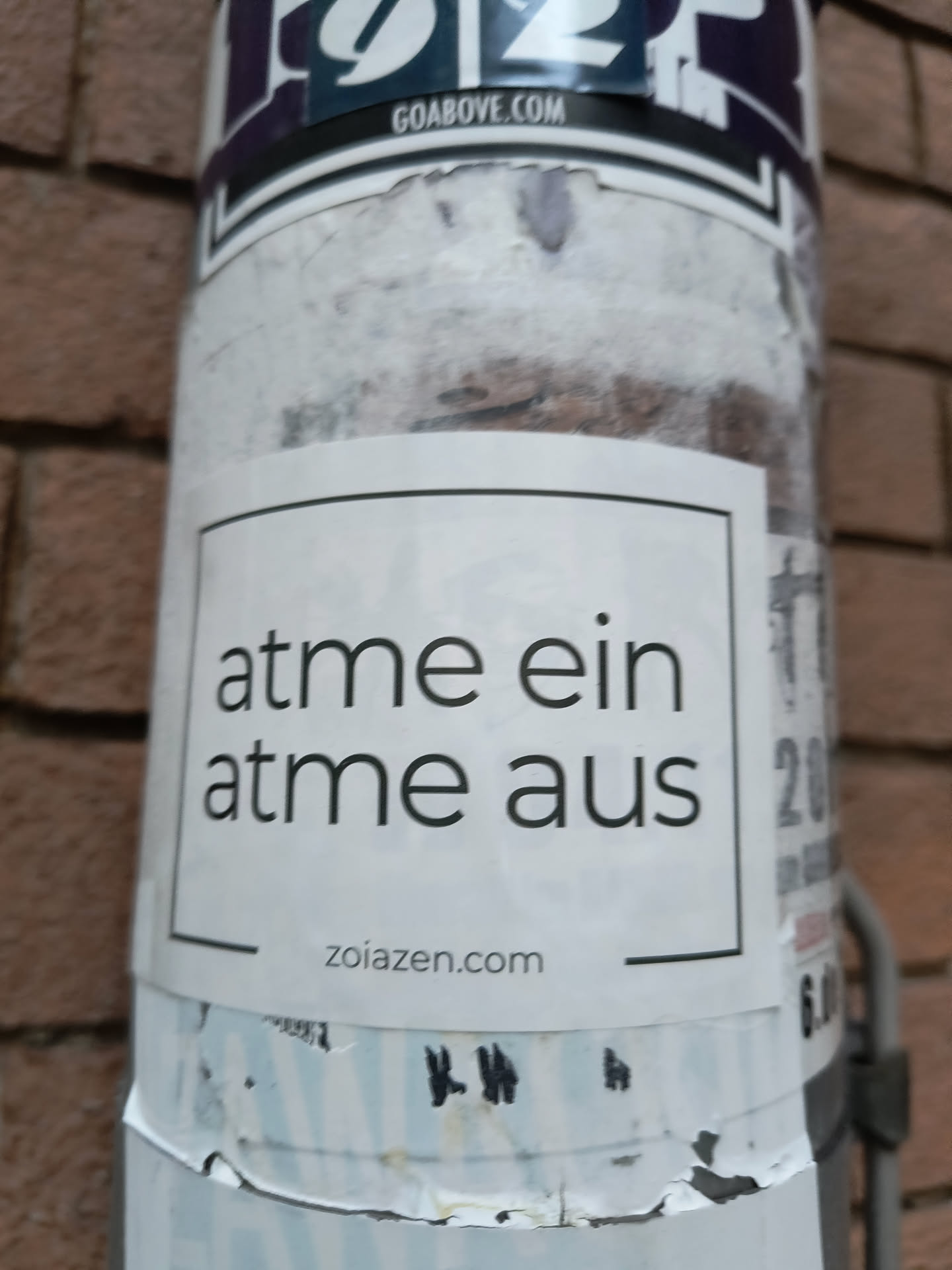 The sticker is white with a black border and contains text in black sans-serif font. The main text reads 'atme ein' on the first line and 'atme aus' on the second line. Below this, centered, is the website address 'zoiazen.com'. The sticker appears to be applied to a cylindrical surface, possibly a pole, and is slightly weathered.