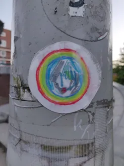 A round sticker with a drawing of a face in the center surrounded by a rainbow. The drawing is done in crayon or colored pencil and appears to be child's artwork. The sticker is affixed to a gray pole, which also has other stickers and markings.