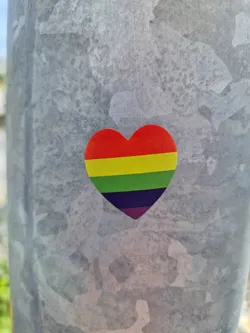 A small, heart-shaped sticker with the rainbow colors of the LGBTQ+ pride flag. It is affixed to a gray metal pole.  The heart is made of vinyl, with a slightly glossy finish.