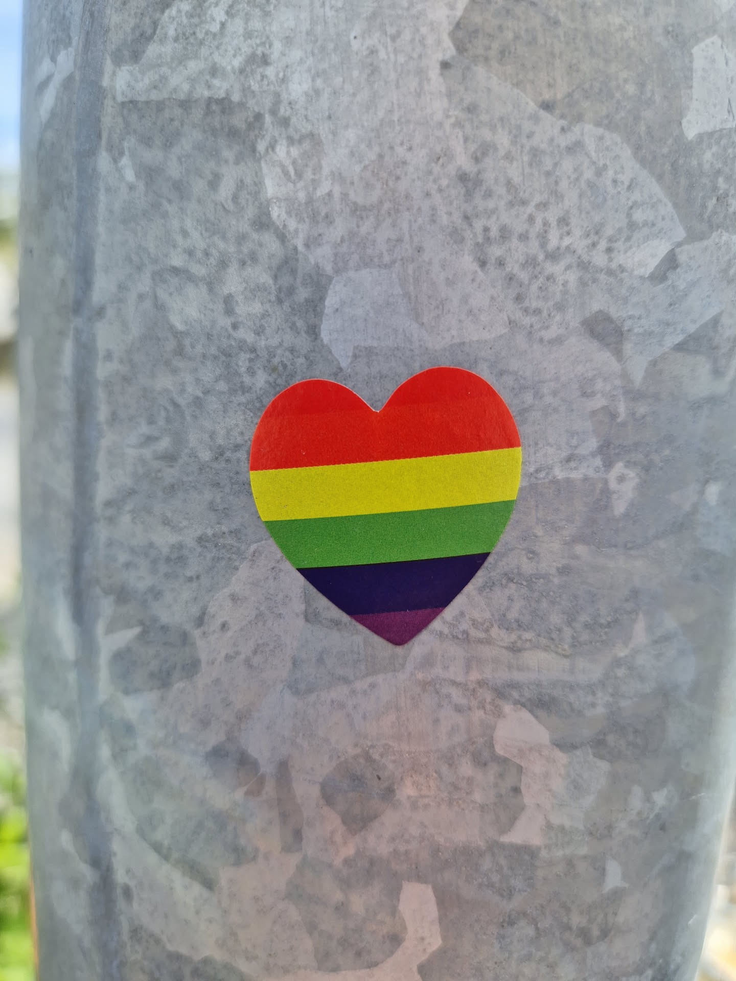 A small, heart-shaped sticker with the rainbow colors of the LGBTQ+ pride flag. It is affixed to a gray metal pole.  The heart is made of vinyl, with a slightly glossy finish.
