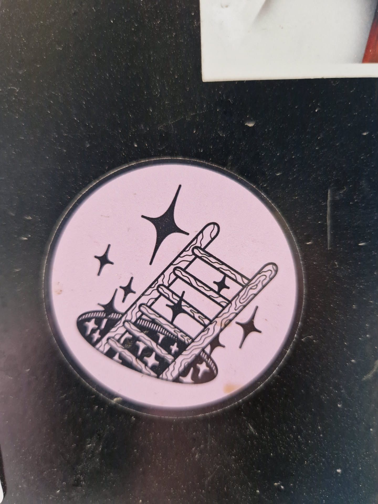 Pink circle sticker with a black line drawing of a ladder falling into a hole. There are small stars surrounding the ladder. 