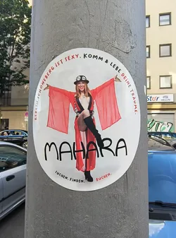 The sticker features an image of a woman in a red robe and a hat, with the words 'BERLIN SCHÖNEBERG IST SEXY. KOMM & LEBE DEINE TRÄUME.' and 'MAHARA' printed on it. The text 'SUCHEN. FINDEN. BUCHEN.' is at the bottom of the sticker.