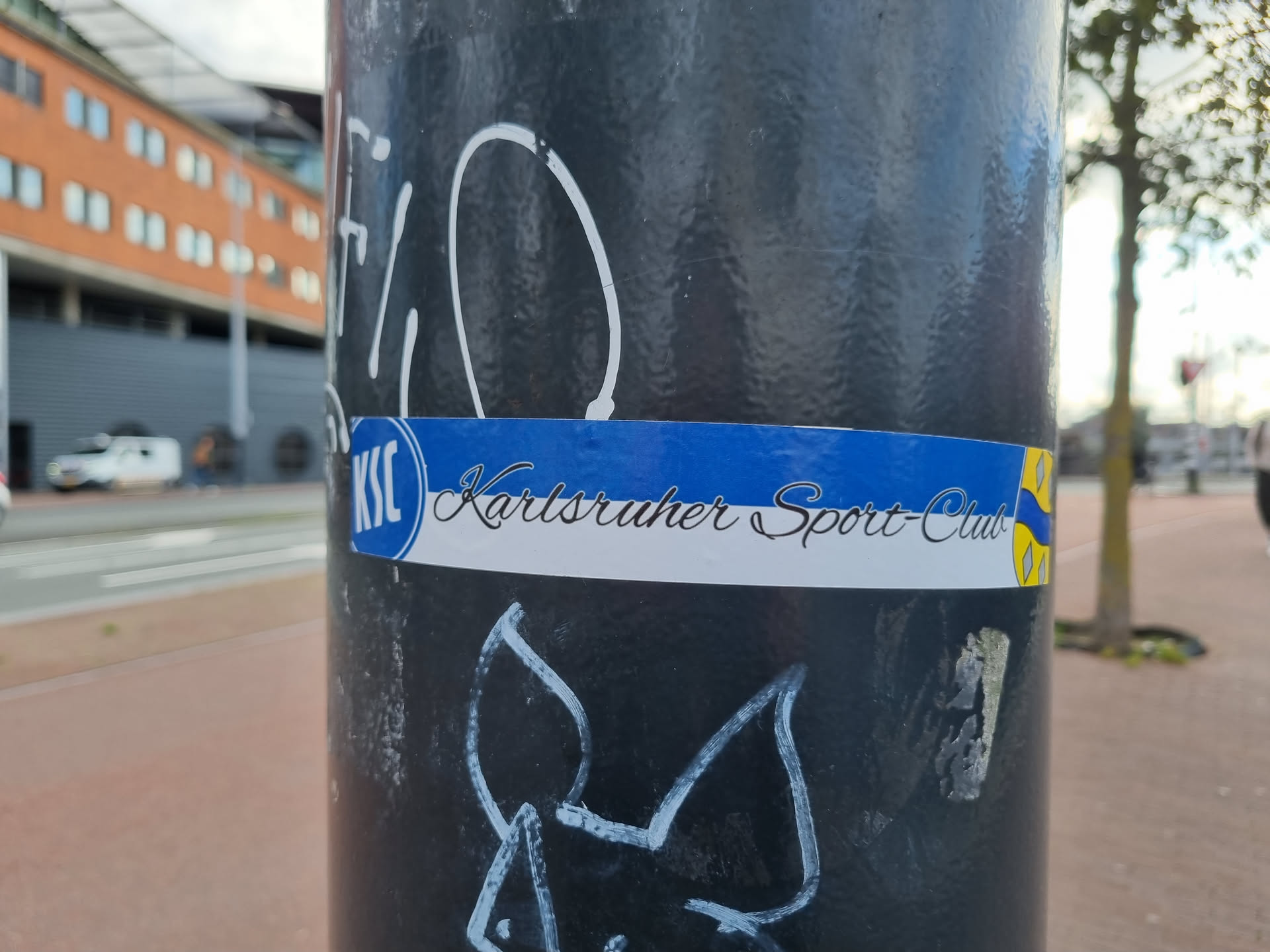 A sticker featuring the logo and name of Karlsruher Sport-Club (KSC), a German football club, affixed to a pole. The sticker is predominantly blue, white, and yellow, with the club's initials and name clearly visible.