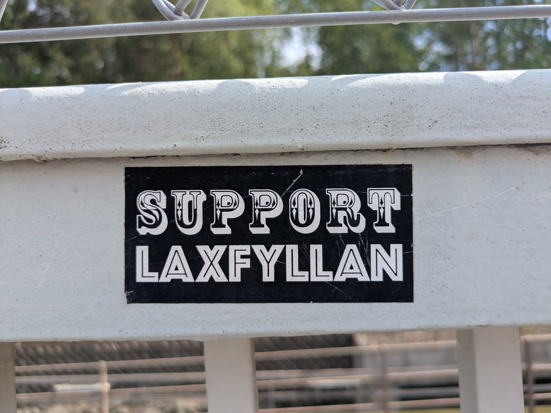 A black rectangular sticker with white text saying "Support Laxfyllan" is adhered to a white, possibly metal, fence or barrier. The text has a decorative font. The sticker looks like it has been there for a while, showing slight wear and tear.