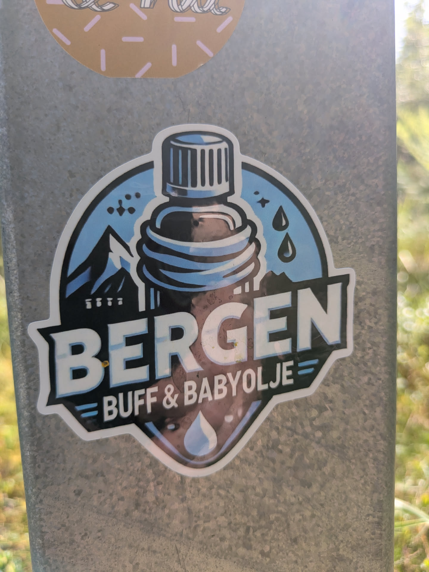 The sticker is shield-shaped with a white border, showcasing a bottle of dark liquid with a blue cap, possibly oil. In the background, there are blue mountains on the left and water droplets on the right against a light blue sky. The bold text 'BERGEN' is prominently displayed in white across the center, with 'BUFF & BABYOLJE' written in smaller letters below it. A single teardrop icon is at the very bottom.