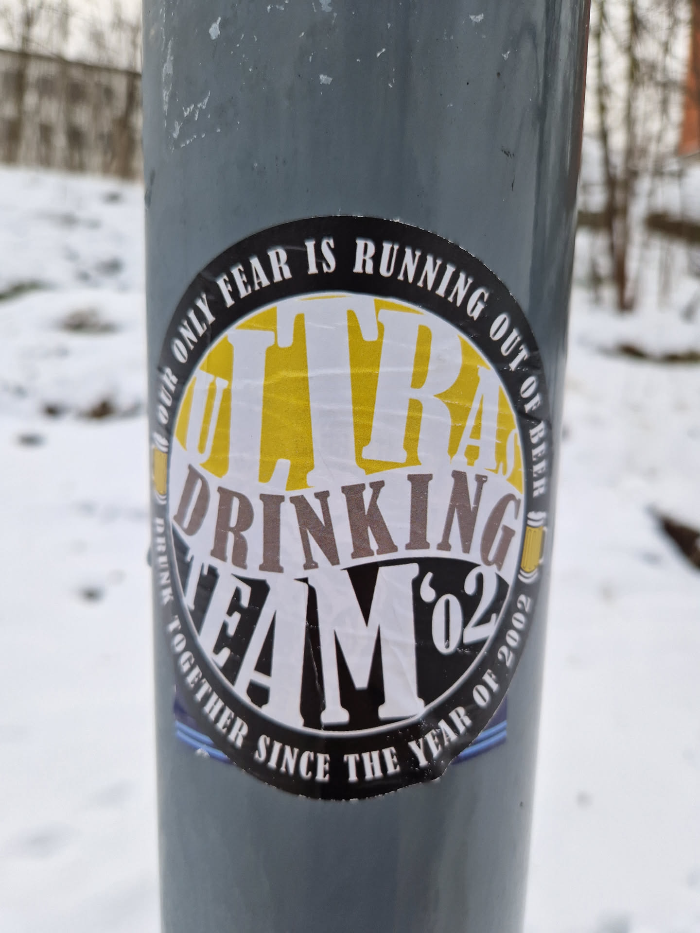 A round sticker with the text "Ultra Drinking Team '02" in the center. The text is in a circle with the words "Our only fear is running out of beer" around it. There is also text at the bottom that says "Together since the year of 2002". The sticker is on a gray pole in a snowy setting.