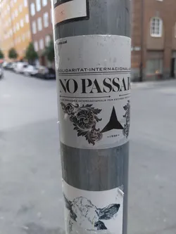 A grayscale sticker with the words "NO PASSARAN" prominently displayed.  The sticker also includes the text "SOLIDARITAT INTERNACIONAL" and what appears to be a year, "1936", suggesting a historical reference. The design features an ornate flourish and a simple geometric shape. It's affixed to a gray metal pole in an urban environment.