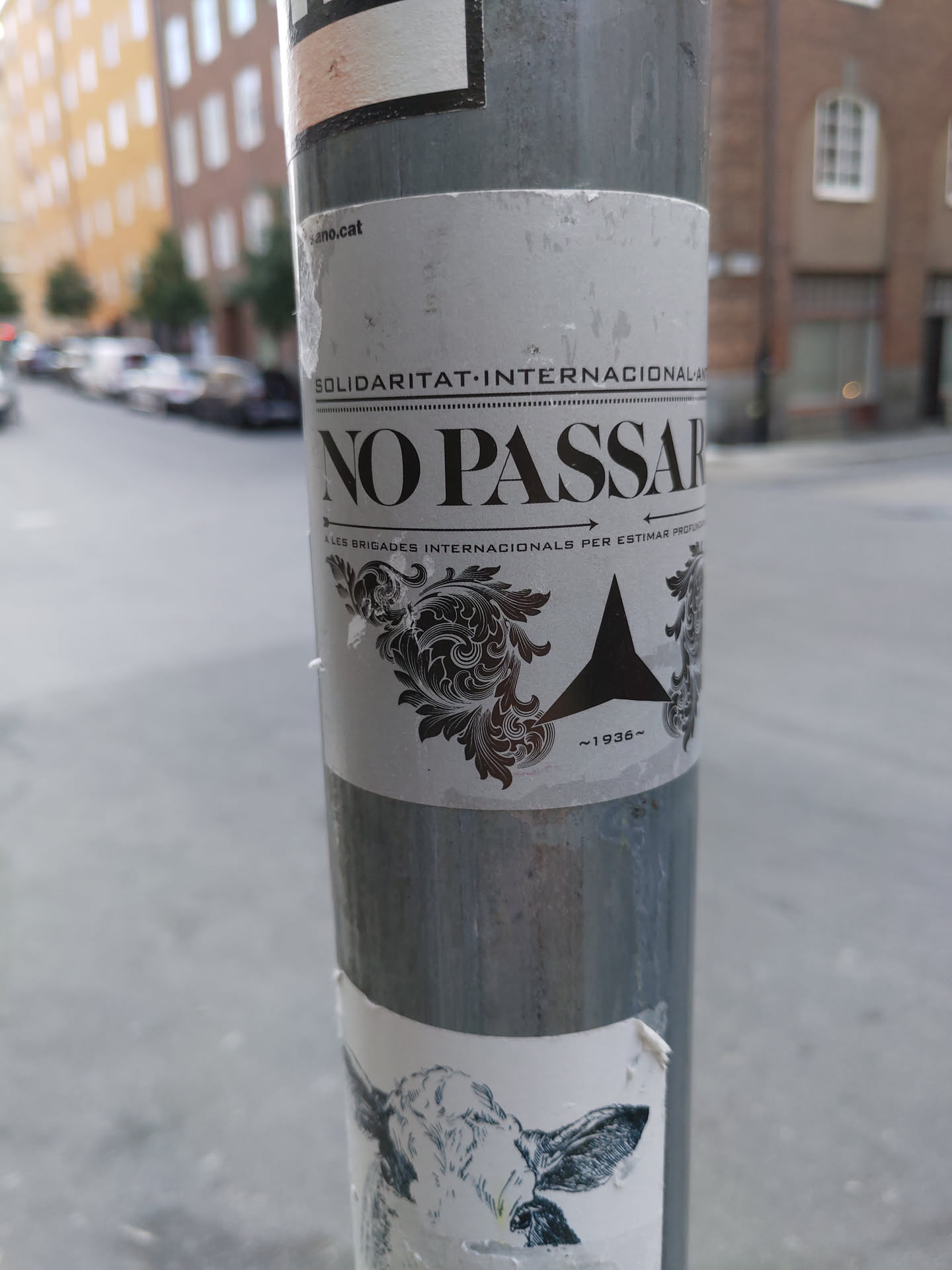 A grayscale sticker with the words "NO PASSARAN" prominently displayed.  The sticker also includes the text "SOLIDARITAT INTERNACIONAL" and what appears to be a year, "1936", suggesting a historical reference. The design features an ornate flourish and a simple geometric shape. It's affixed to a gray metal pole in an urban environment.