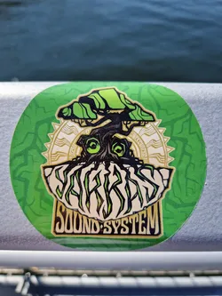 This is a round sticker featuring vibrant green and gold artwork. The central image depicts a stylized tree with a face, seemingly emerging from a rock formation.  The text "Yak Bass Sound System" is prominently displayed beneath the tree. The design aesthetic is a blend of psychedelic and nature-inspired elements.