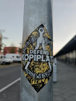 A diamond-shaped sticker on a metal pole.  The sticker features black and yellow imagery with the text "DEFEND KOPIPLATZ" prominently displayed, along with the phrases "KOPIBLEIBT" and "KOPINEWS BERLIN." There are also several smaller illustrations and symbols surrounding the central text. The style is reminiscent of protest or activist art.