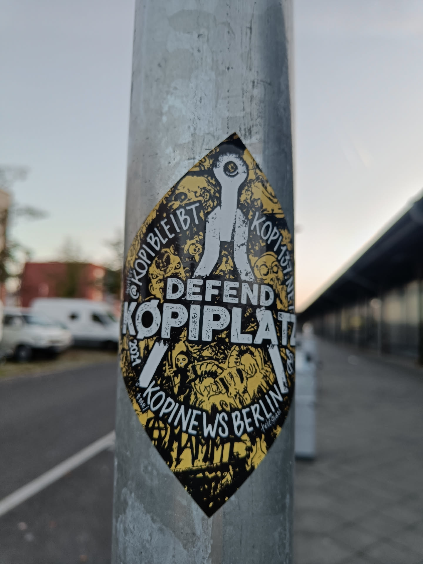 A diamond-shaped sticker on a metal pole.  The sticker features black and yellow imagery with the text "DEFEND KOPIPLATZ" prominently displayed, along with the phrases "KOPIBLEIBT" and "KOPINEWS BERLIN." There are also several smaller illustrations and symbols surrounding the central text. The style is reminiscent of protest or activist art.