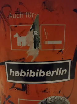 A black and white sticker with the text "Habibi Berlin" on an orange surface. The sticker is rectangular with white lettering on a black background.  There's other graffiti and stickers on the surface as well.
