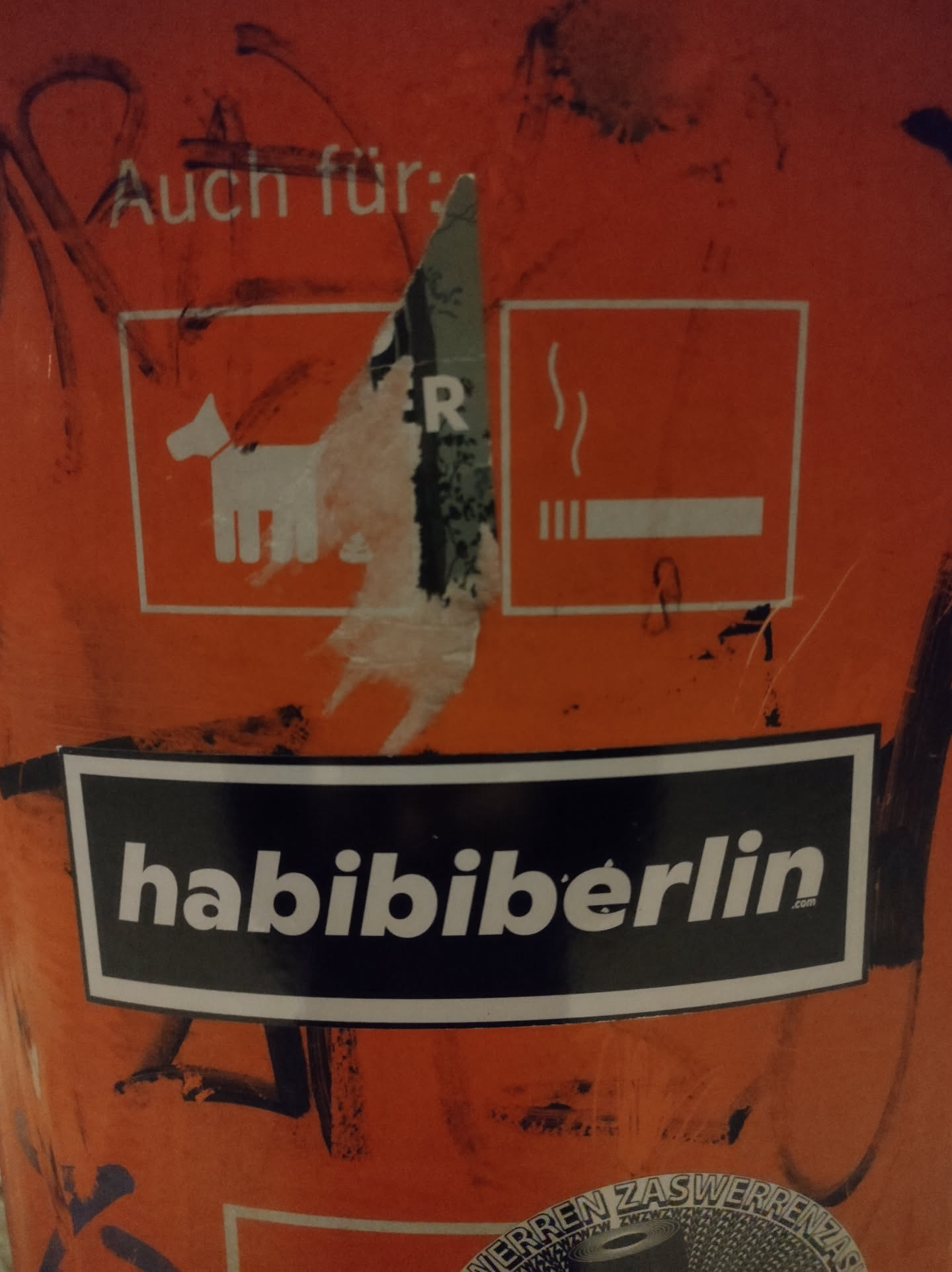A black and white sticker with the text "Habibi Berlin" on an orange surface. The sticker is rectangular with white lettering on a black background.  There's other graffiti and stickers on the surface as well.