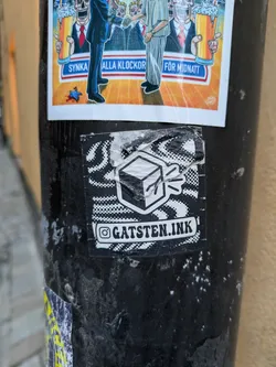 A black and white sticker with a cube design and the text "@gatsten.ink". The sticker is placed on a dark-colored pole with other stickers.