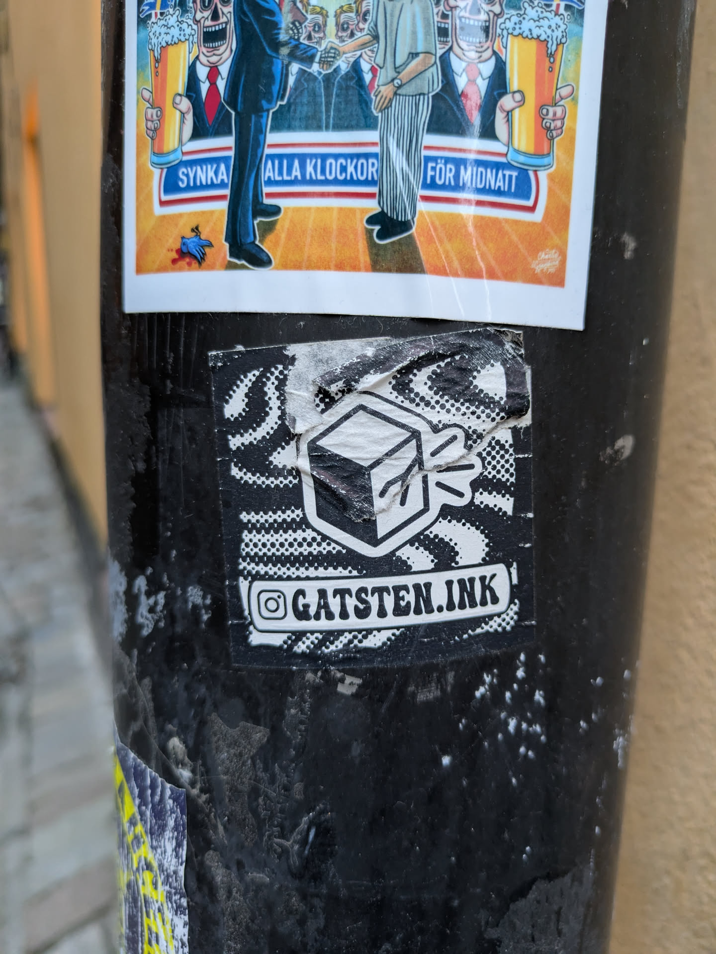 A black and white sticker with a cube design and the text "@gatsten.ink". The sticker is placed on a dark-colored pole with other stickers.