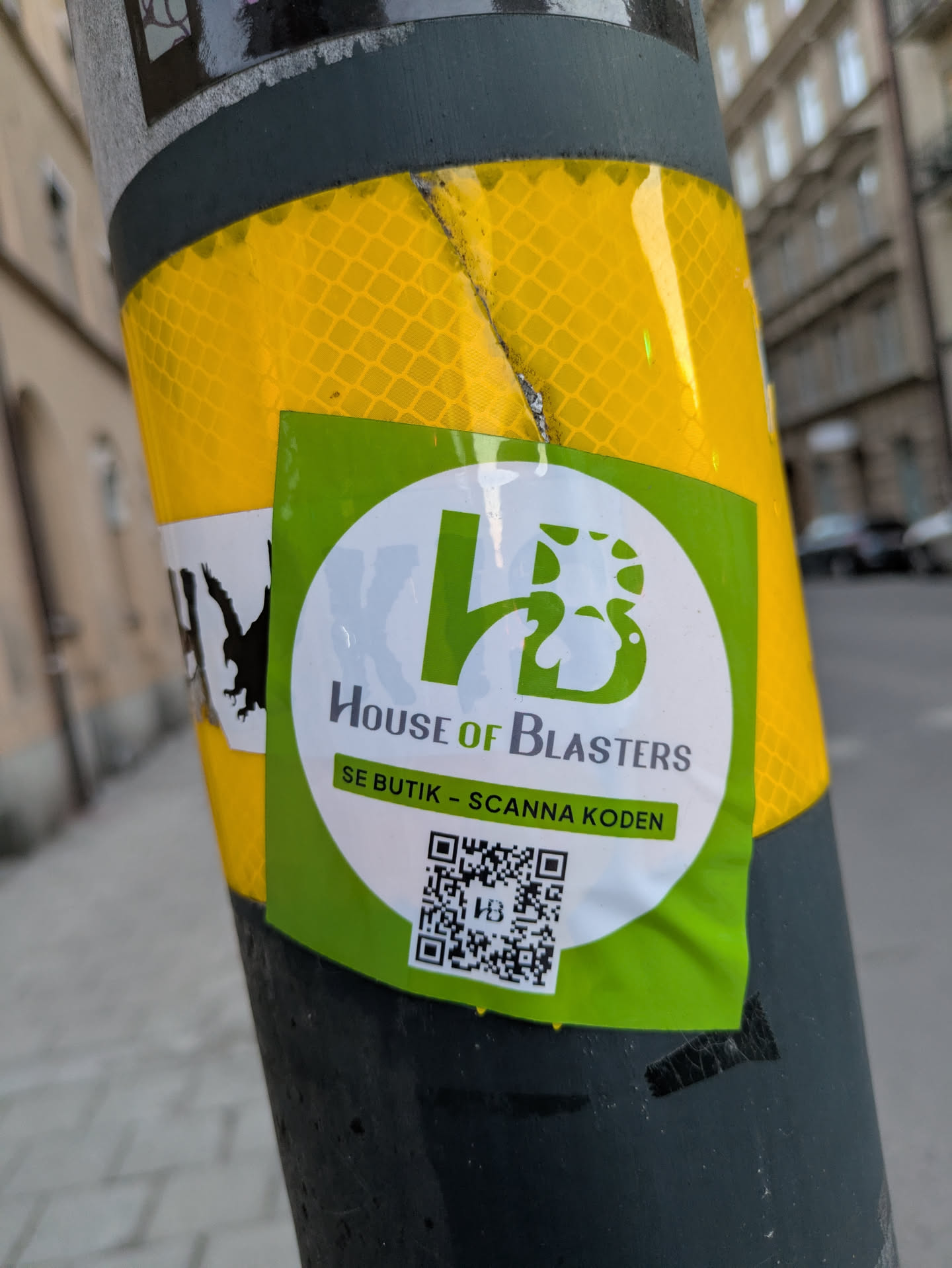 A green and white sticker is affixed to a pole. The sticker features the logo of "House of Blasters" and contains a QR code with the text "SE BUTIK - SCANNA KODEN". The sticker invites people to scan the code to visit the store.
