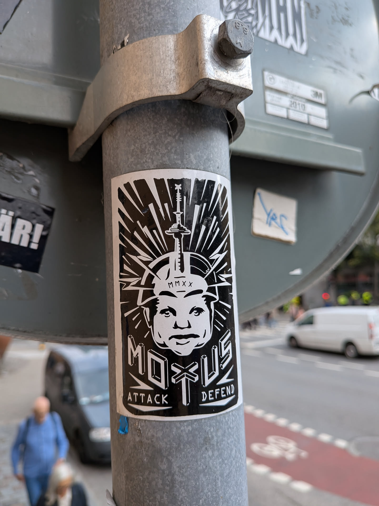 The sticker is predominantly black and white. It features a stylized portrait of a person's head, with a sharp, angular design. A sword or similar pointed object is positioned vertically on top of the head. The words 'MOTUS' and 'ATTACK DEFEND' are prominently displayed. The background behind the face has radiating lines, giving it a dynamic and attention-grabbing appearance. The overall style is graphic and somewhat aggressive.