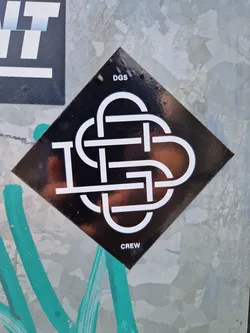 A black diamond-shaped sticker with a white logo. The logo is a stylized knot or intertwined design with the letters DGS incorporated.  The words "DGS" and "CREW" are printed above and below the logo respectively.