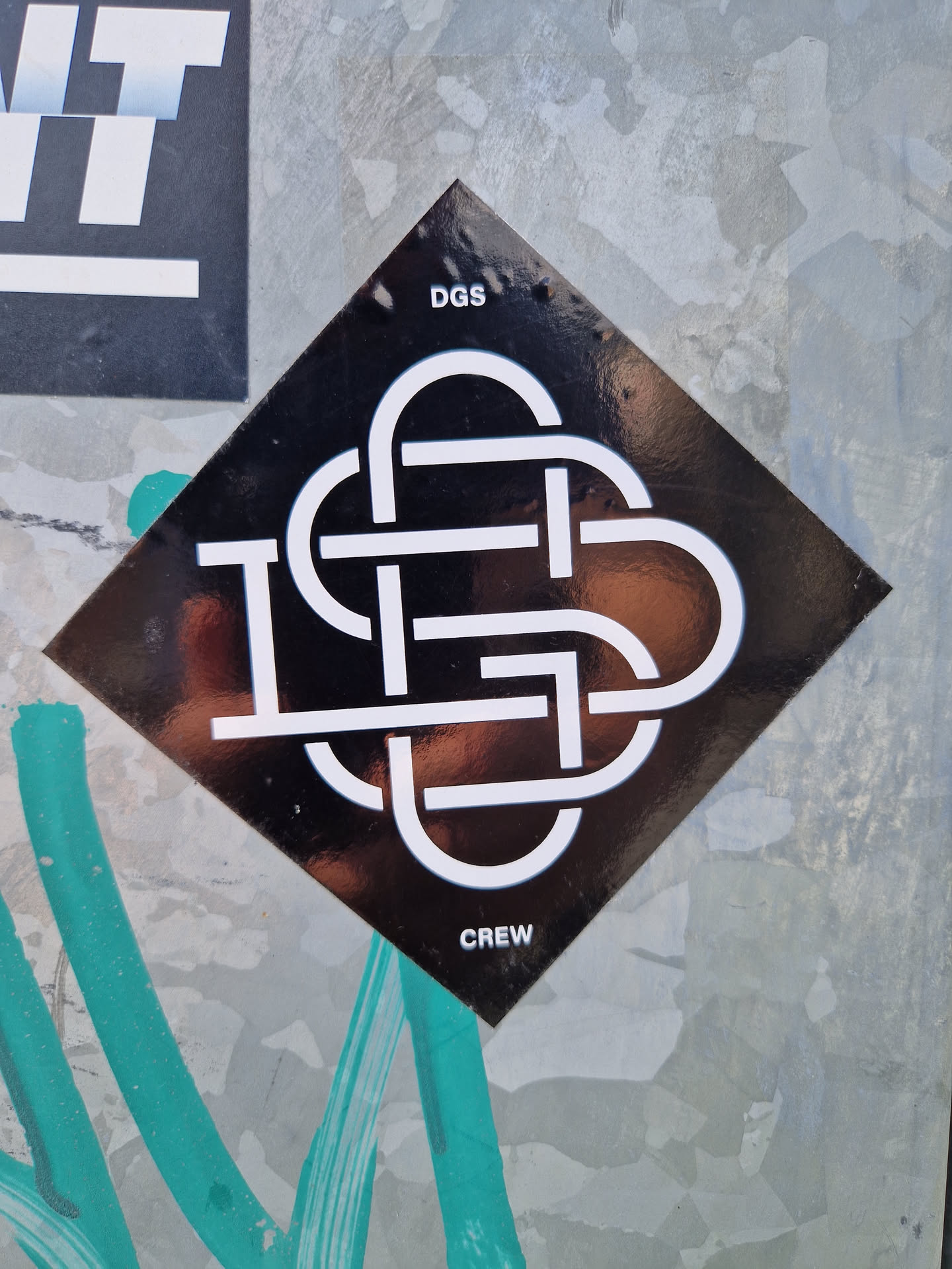 A black diamond-shaped sticker with a white logo. The logo is a stylized knot or intertwined design with the letters DGS incorporated.  The words "DGS" and "CREW" are printed above and below the logo respectively.