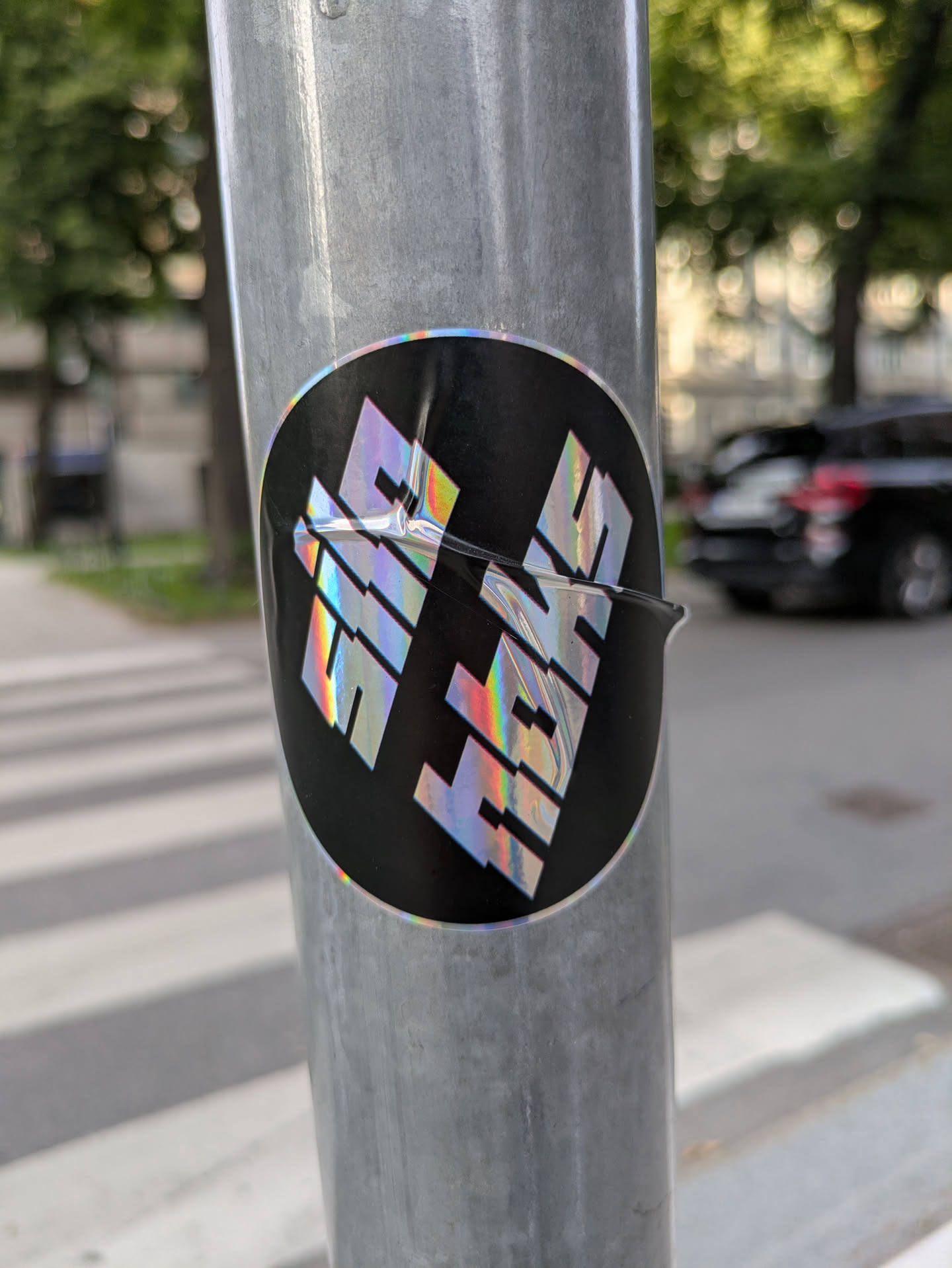 The sticker is circular with a black background. Centered on the sticker are the letters 'HIVE' rendered in a bold, blocky, geometric font. The letters are a striking holographic silver that refracts light, displaying a spectrum of rainbow colors. The sticker appears to be slightly creased or damaged in the center, affecting the top portion of the 'H' and 'I'.