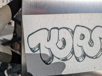 The sticker is white with a distressed, cracked surface. It features bold, black bubble letters that form a word or tag. The letters have thick outlines and some internal detailing, such as parallel lines and small circles, giving them a stylized, graffiti-like appearance. The overall impression is one of age and exposure to the elements.
