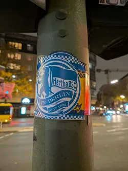 Street sticker A rectangular fan sticker adhered to a metal street pole at night. It features a blue and white circular emblem depicting the Berlin Bear holding a Hertha BSC flag. Below the flag are banners inscribed with 'IN BERLIN' and 'SEIT 1892'. The top and bottom edges are lined with blue-and-white checkered patterns, flanked by yellow graphic accents on the sides.
