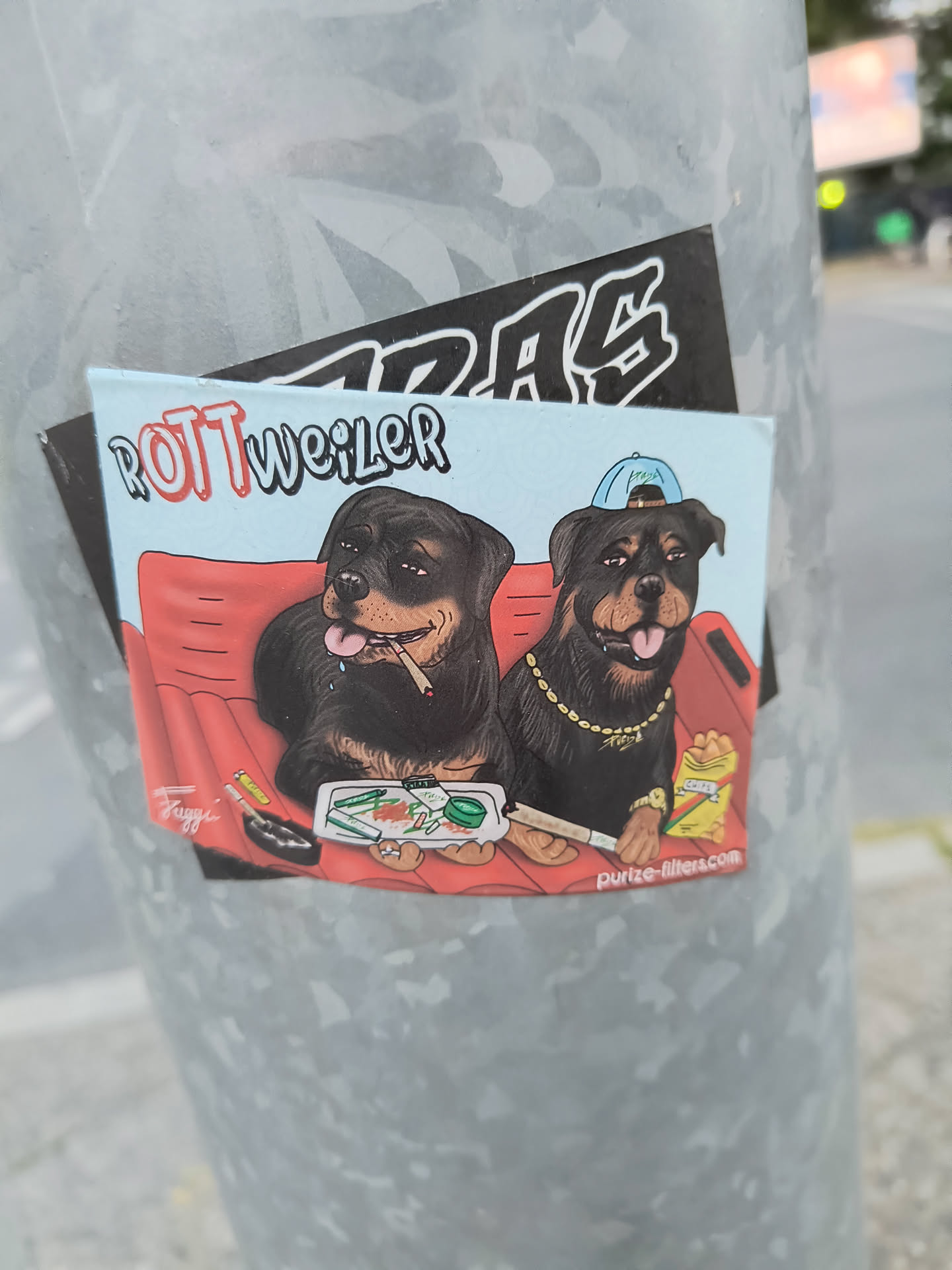 The sticker features a cartoon illustration of two Rottweiler dogs sitting on a red couch against a light blue background. The dog on the left is smoking a cigarette, with smoke curling from its mouth. The dog on the right wears a blue baseball cap, a gold chain, and a wristwatch. Both dogs appear relaxed. In front of them is a tray with rolling papers, a grinder, and filters, suggesting they are preparing to smoke. There is also a bag of chips and what looks like an ashtray. The text 'Rottweiler' is prominently displayed above the dogs. The overall style is cartoonish with bold outlines and bright colors.