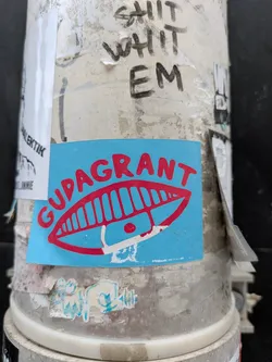 The sticker is light blue and has "GUDAGRANT" written in red, with a stylized red graphic that looks like a mouth with teeth above the text. The sticker is affixed to a light-colored, weathered surface.