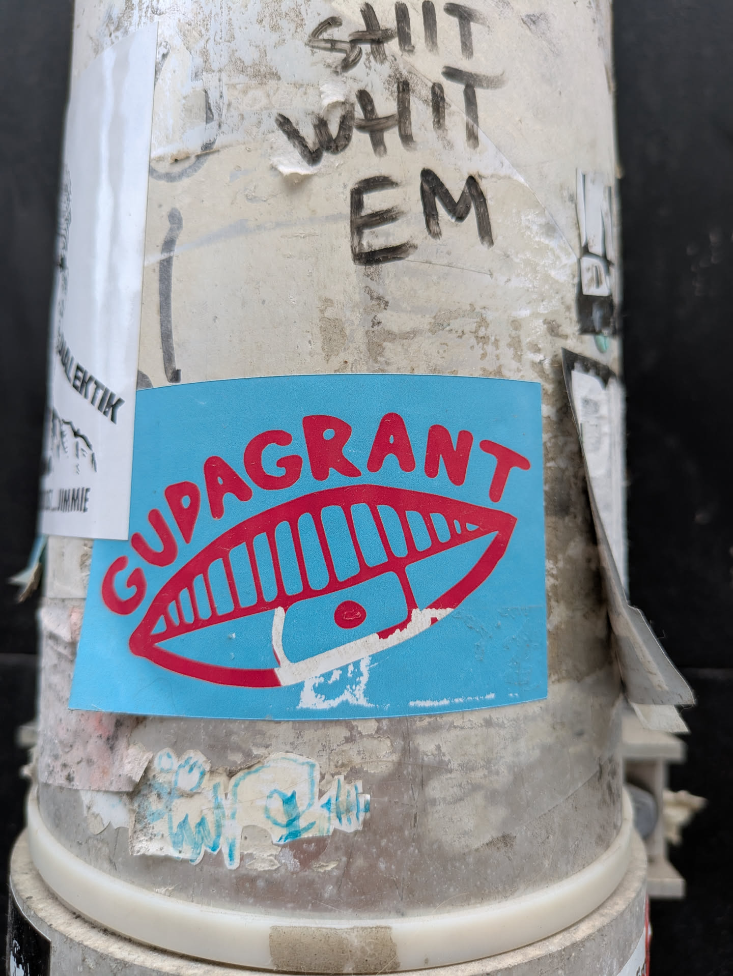 The sticker is light blue and has "GUDAGRANT" written in red, with a stylized red graphic that looks like a mouth with teeth above the text. The sticker is affixed to a light-colored, weathered surface.