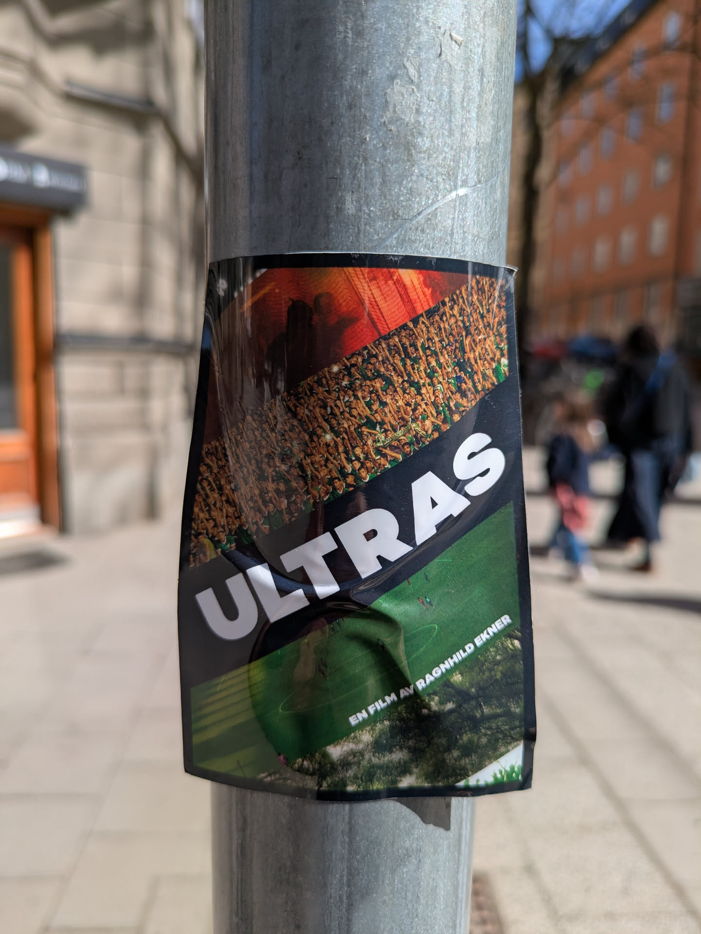 This sticker is a poster for the movie "Ultras." The top of the sticker shows a crowd of people in red and orange. The word "ULTRAS" is printed in white letters diagonally across the image. The bottom section shows a view of a soccer field. The sticker is affixed to a pole.