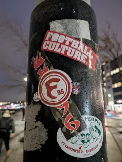 A collection of stickers on a pole, the main one is round with the letter F and the number 95. There is also a sticker that says Football Culture and another with a camouflage pattern and some text in a different language.
