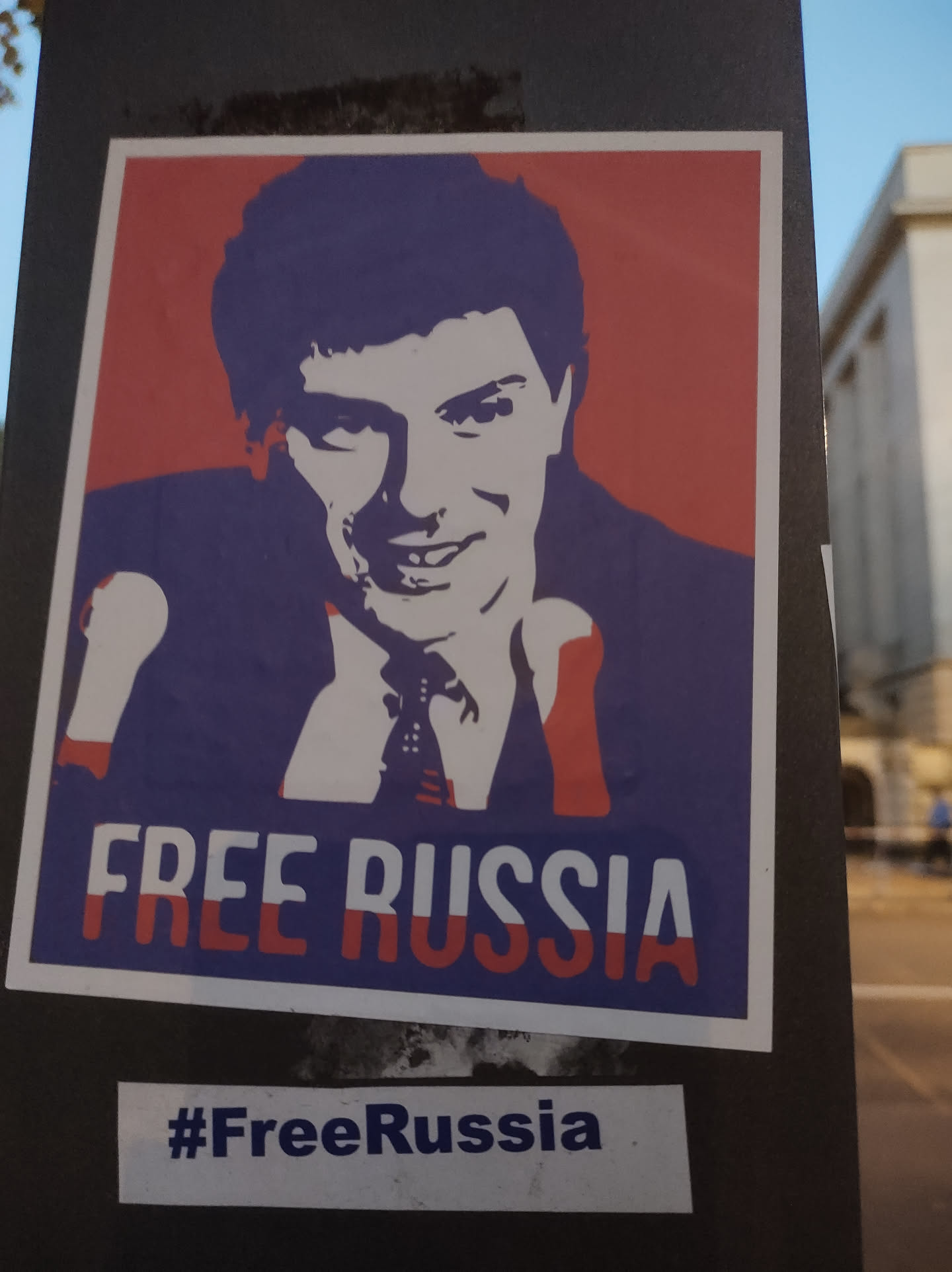 A sticker featuring a stylized portrait of a man, reminiscent of Soviet-era propaganda posters, with the text "FREE RUSSIA" prominently displayed. The colors are primarily red, white, and blue, evoking the Russian flag. A smaller sticker below reads "#FreeRussia".