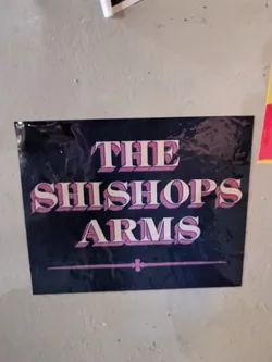 A sticker featuring the text 'The Shishopps Arms' in a retro-style font. The text is pink on a dark background, with a thin line separating the name and 'Arms'.