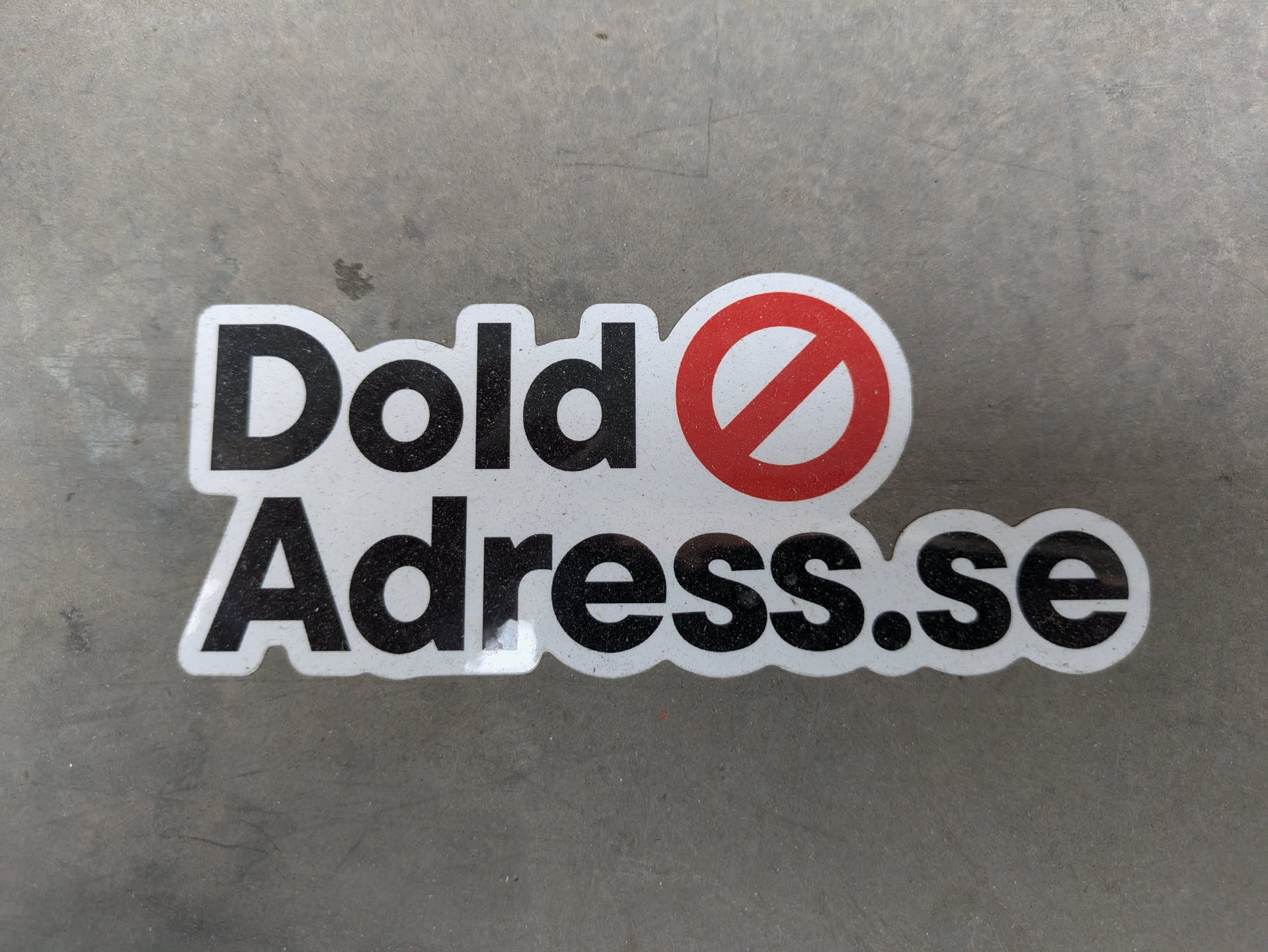 The sticker is white with black text that reads "Dold Adress.se". There is a red circle with a line through it, replacing the letter 'O' in 'Dold'. The background is gray.