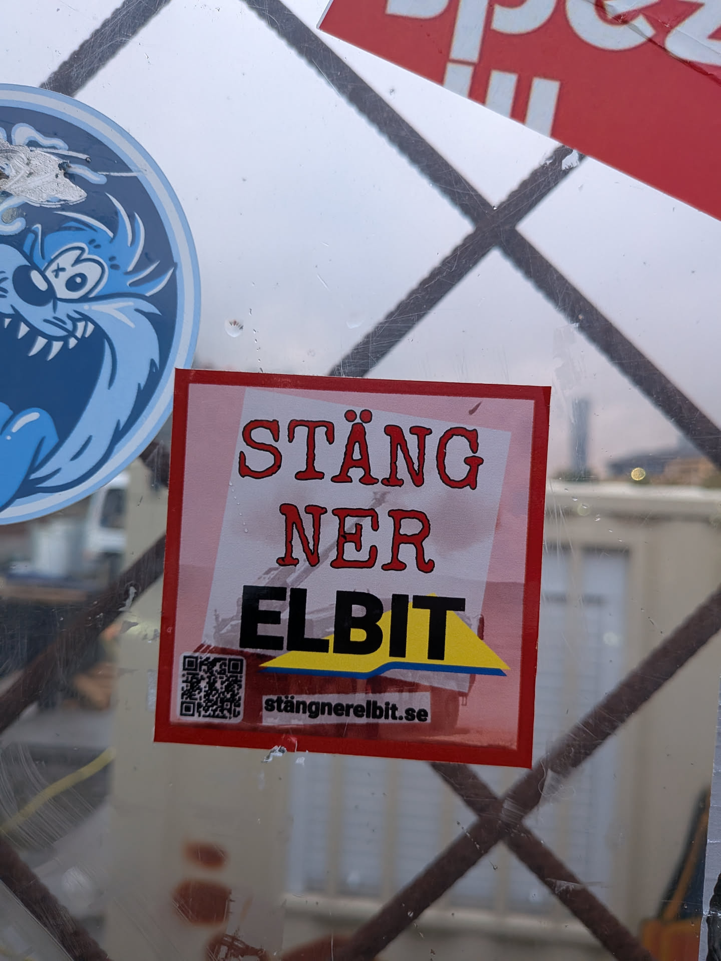 The sticker is square-shaped with a red border. The background is a gradient of pink and white. The text 'STÄNG NER' is written in a stylized, bold red font. Below this, 'ELBIT' is written in large, bold black letters, with a yellow and blue graphic element underneath. A QR code is present on the left side of the sticker, and the website 'stängnerelbit.se' is printed below it. The sticker appears to be adhered to a reflective surface, possibly glass, with some scuff marks and reflections visible.