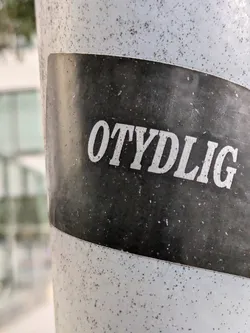 A black and white sticker with the word "OTYDLIG" in a bold, sans-serif font. The sticker is affixed to a speckled grey pole. 
