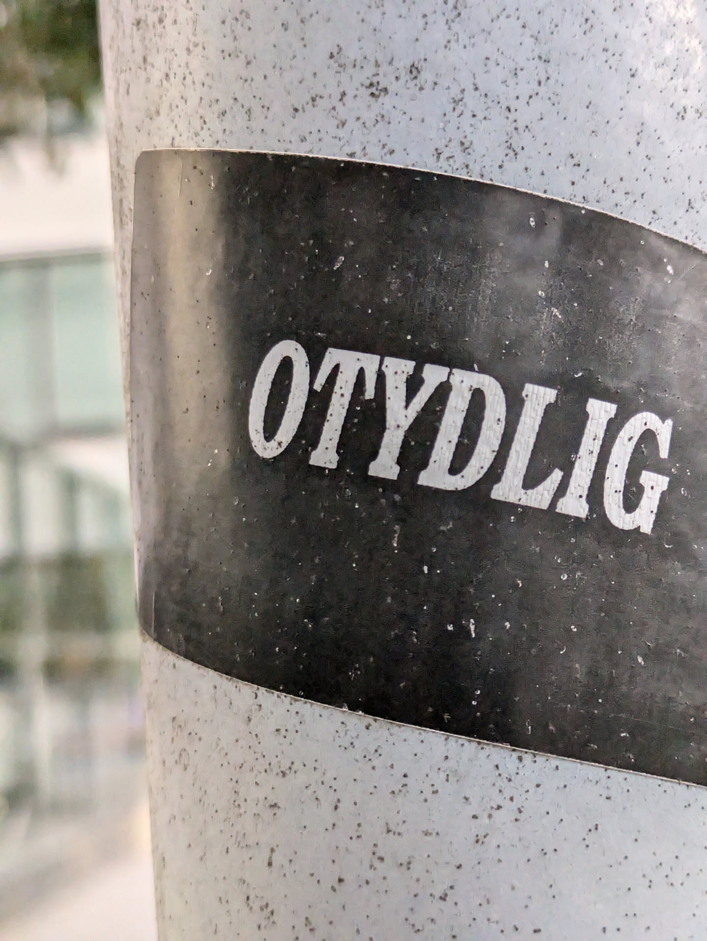 A black and white sticker with the word "OTYDLIG" in a bold, sans-serif font. The sticker is affixed to a speckled grey pole. 