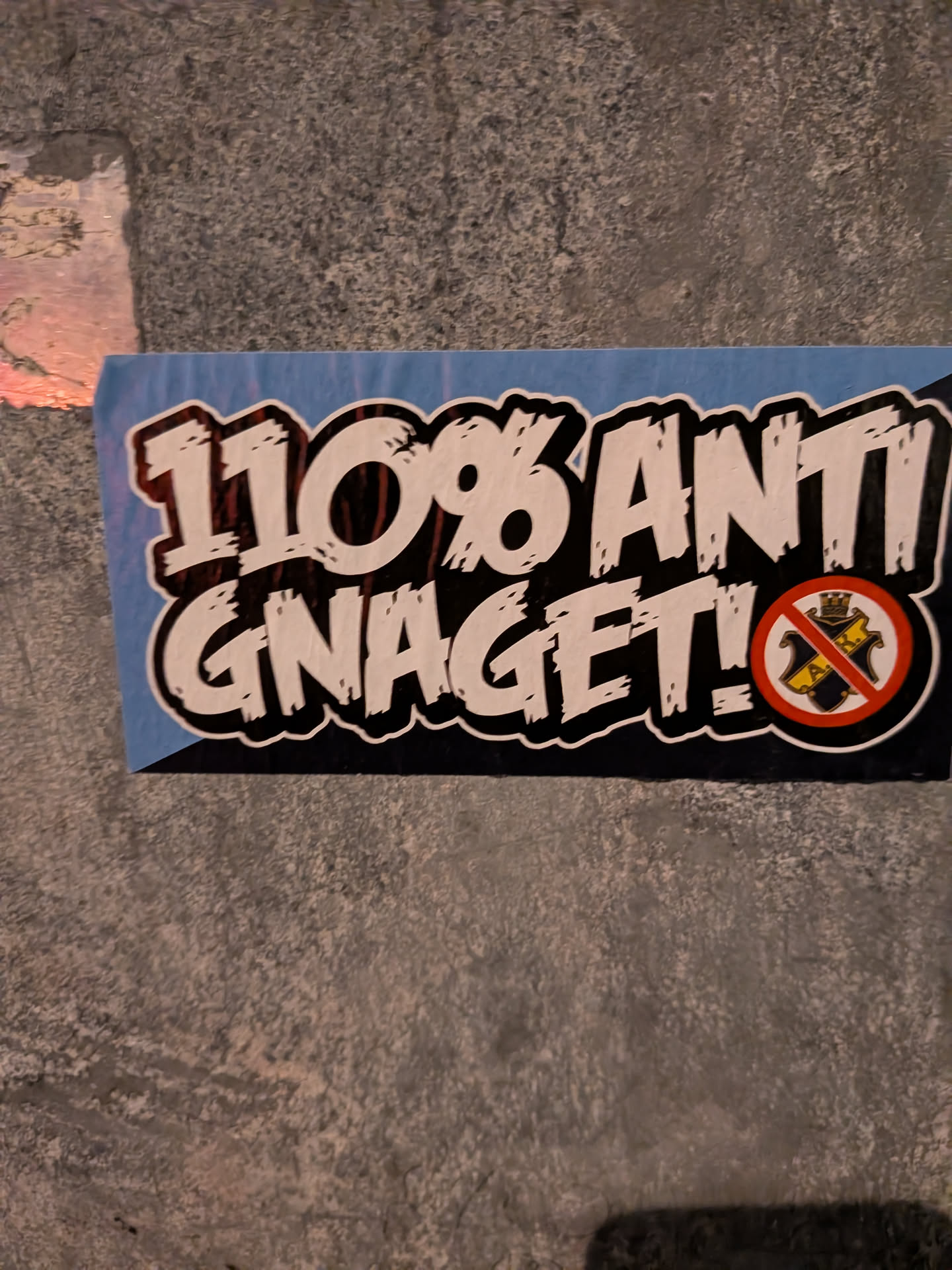 A rectangular street sticker featuring bold, distressed white lettering with thick black outlines that reads '110% ANTI GNAGET!'. The background features light blue and dark blue sections. On the lower right, the crest of AIK (Allmänna Idrottsklubben) is displayed inside a red prohibition circle with a diagonal red line crossing it out.