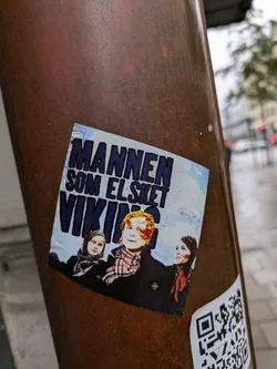 A sticker featuring the movie poster of Mannen som elsket viking (The Man Who Loved Viking).  The poster shows three people, two women and one man with red hair, against a snowy backdrop. A QR code is present on the sticker.