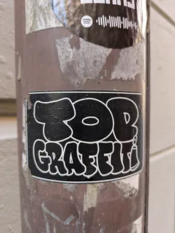 A black and white sticker with the text "TOB GRAFFITI" in a bubbly font, adhered to a brown metal pole. There is another sticker above it with a Spotify logo.