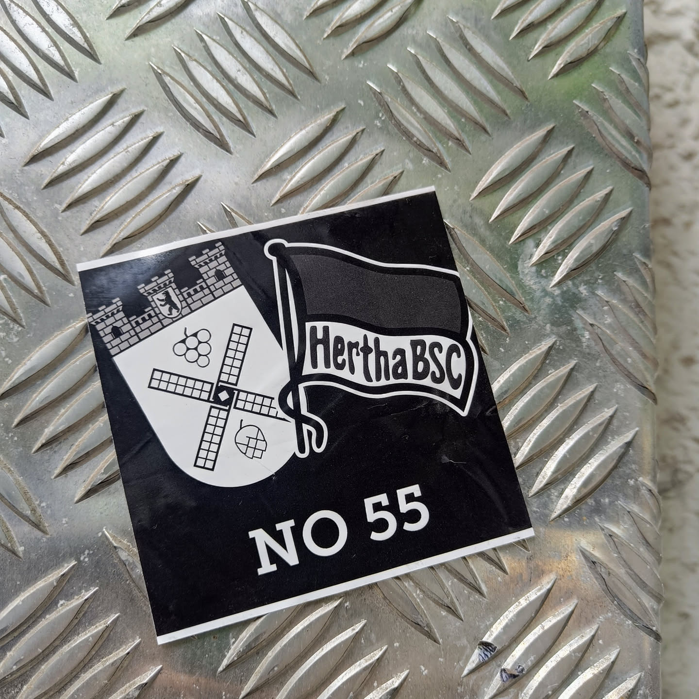 Black and white sticker featuring the Hertha BSC logo and the number 55. The logo includes a coat of arms with a windmill and grapes, and a flag with the club's name.  The sticker is affixed to a metal surface.