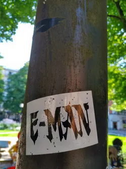 A white sticker with the text "E-MAN" in black stylized lettering, stuck on a dark brown pole in an outdoor setting. The lettering style is sharp and edgy.
