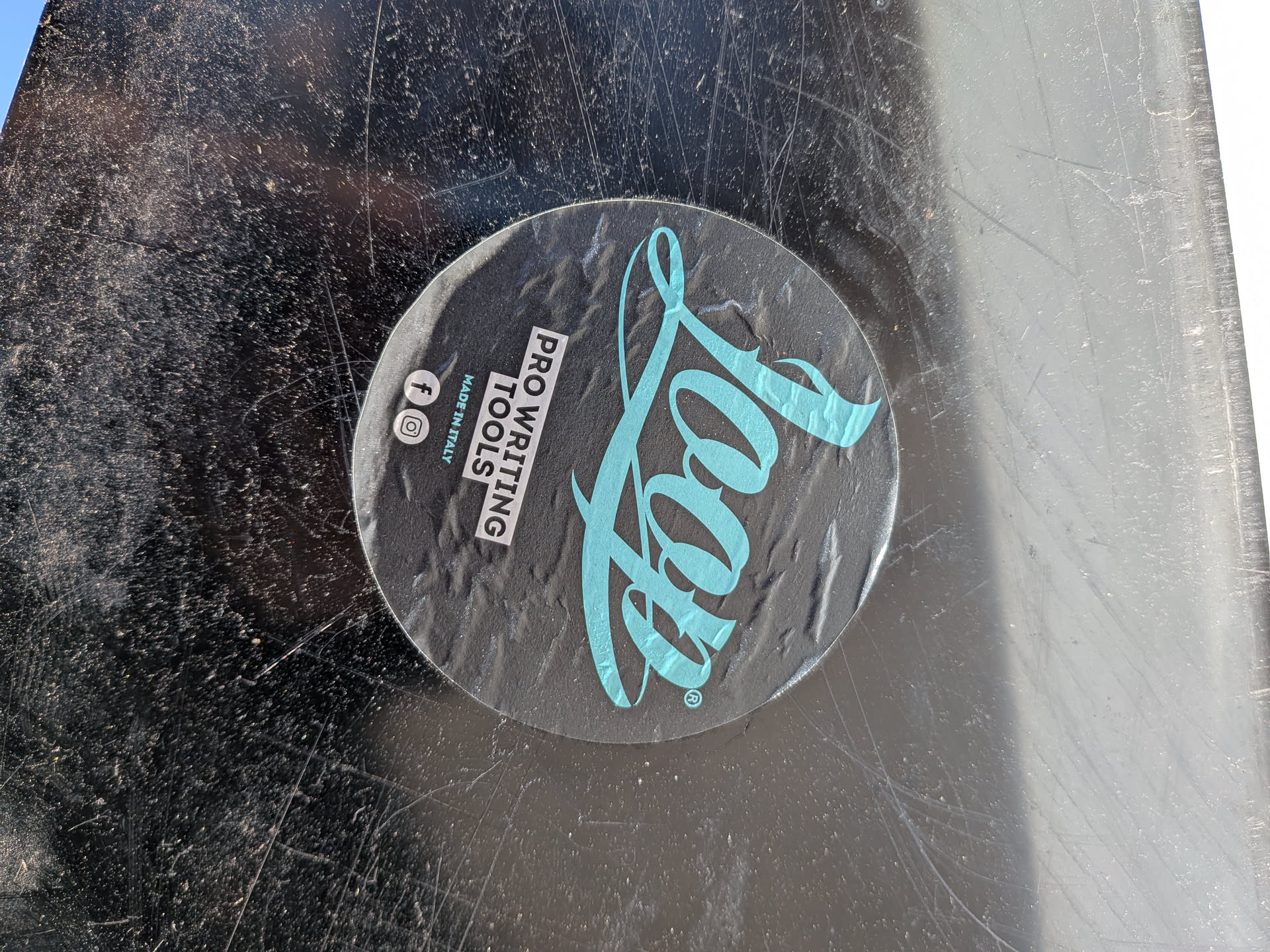 A circular sticker with a dark grey background. The prominent feature is a large, stylized 'Loop' logo in a vibrant light blue/teal color, positioned on the right side with a registered trademark symbol. On the left side, stacked vertically in white sans-serif text, it reads 'PRO WRITING TOOLS' followed by 'MADE IN ITALY'. Below this text, there are two white social media icons: Facebook and Instagram. The sticker appears slightly worn and wrinkled, applied to a heavily scratched, dark surface.