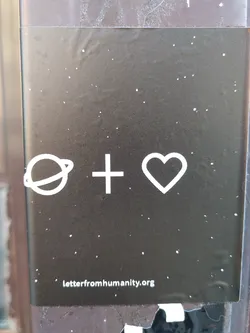 A dark-colored sticker with white symbols: a planet, a cross and a heart.  The text 'letterfromhumanity.org' is visible at the bottom.
