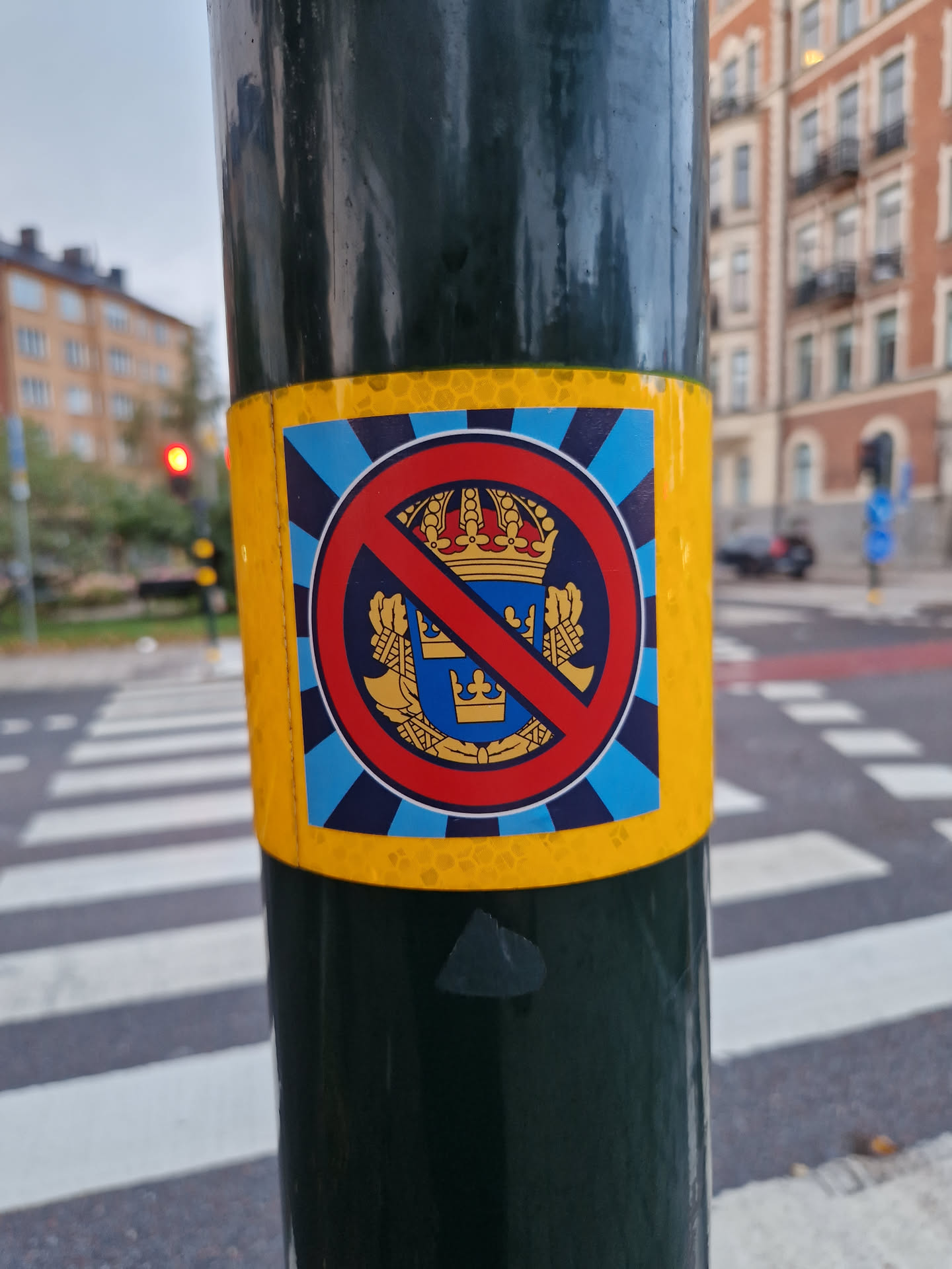 A sticker featuring a red circle-slash symbol over a coat of arms, likely representing opposition to monarchy.  The sticker has a blue and yellow color scheme and is affixed to a pole.