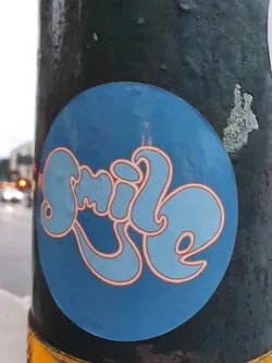 A round blue sticker with the word "Smile" written in a stylized, groovy font. The letters are light blue with an orange outline, creating a retro feel. The sticker is affixed to a dark-colored pole.