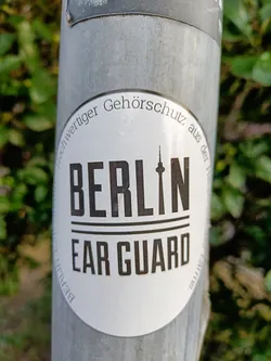 A round sticker with the text "BERLIN" and "EAR GUARD" in black text on a white background.  A small silhouette of the Berlin Television Tower is visible between the two lines of text. The circular border has the words  "Hochwertiger Gehörschutz aus der Hauptstadt Berlin" which translates to "High-quality hearing protection from the capital Berlin".