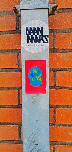 A square sticker with a red background features a pixelated image resembling a thermal scan of a head. The colors range from blue to green and red, indicating different temperature levels. The sticker is affixed to a metal pole.